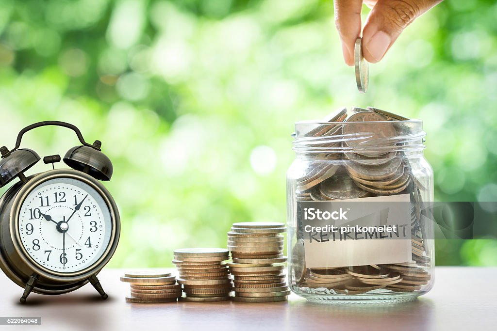 Hand putting Coins in glass jar with retro alarm clock  for time to money saving for retirement concept